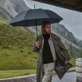 Mann hält einen Knirps Timber Stockschirm in der Hand und steht in einer Bergregion, im Hintergrund sind Berge zu sehen.