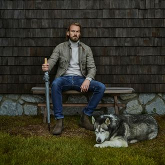 Mann sitzt mit Regenschirm auf einer Parkbank, neben ihm liegt ein Husky-Hund und wartet ruhig im Regen.