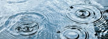 Regentropfen fällt auf Wasseroberfläche und erzeugt kreisförmige Wellen